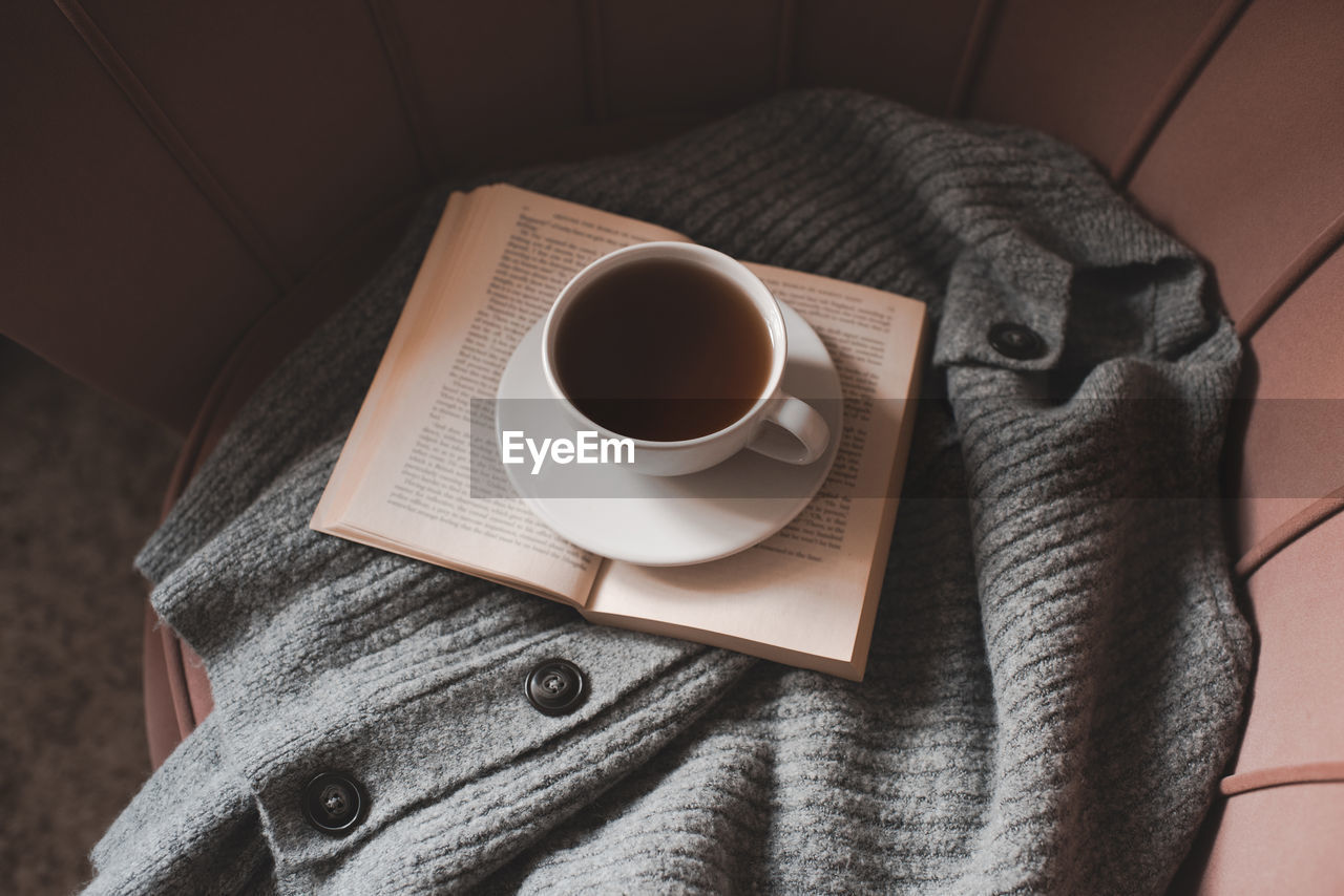 Mug of coffee with open paper book, knitted textile on cozy armchair at home closeup. good morning.