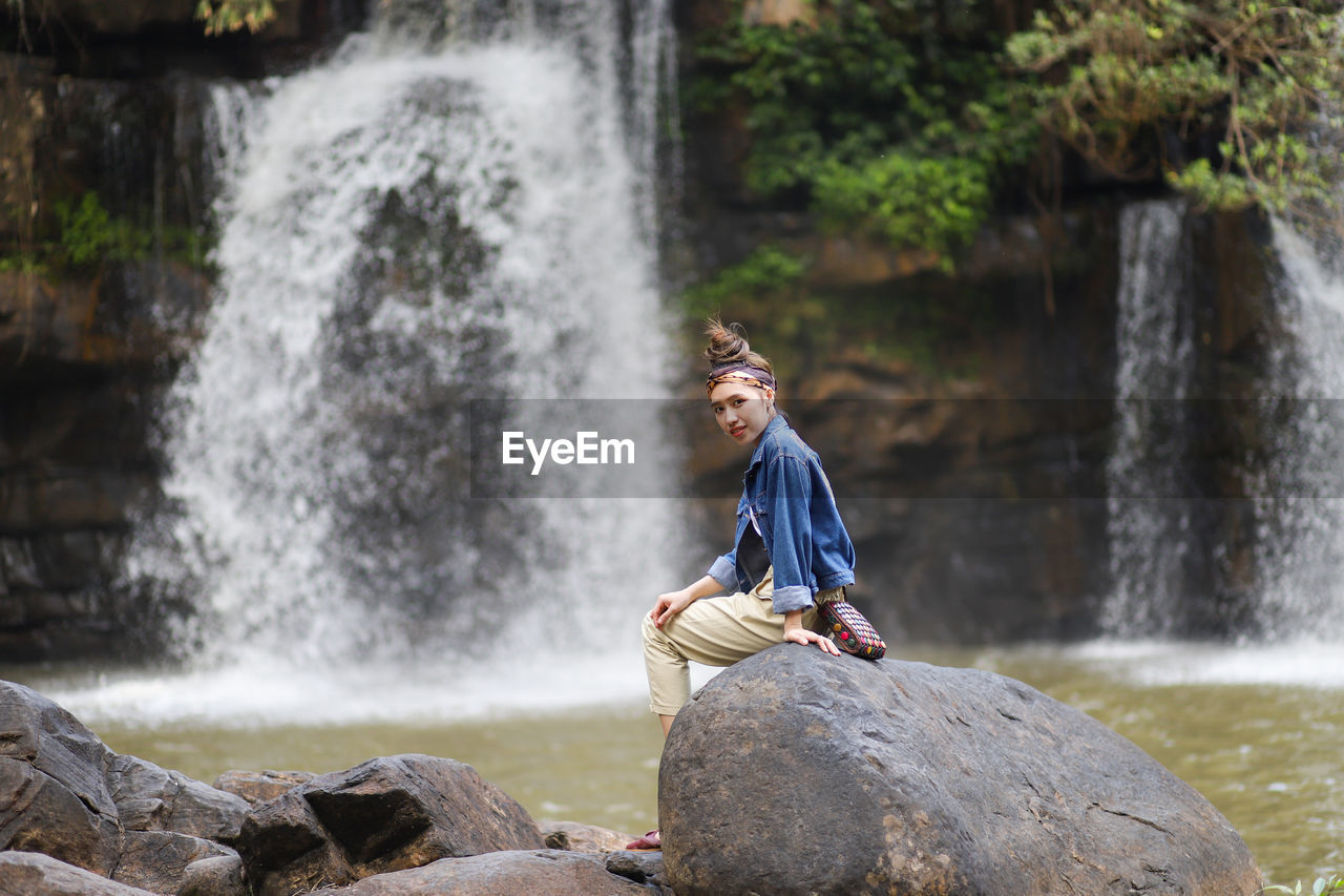 Portrait of smiling woman sitting on rock against waterfall