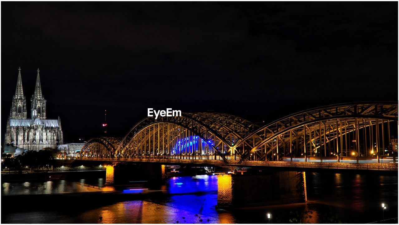 Illuminated hohenzollern bridge over rhine river by cologne cathedral against sky at night