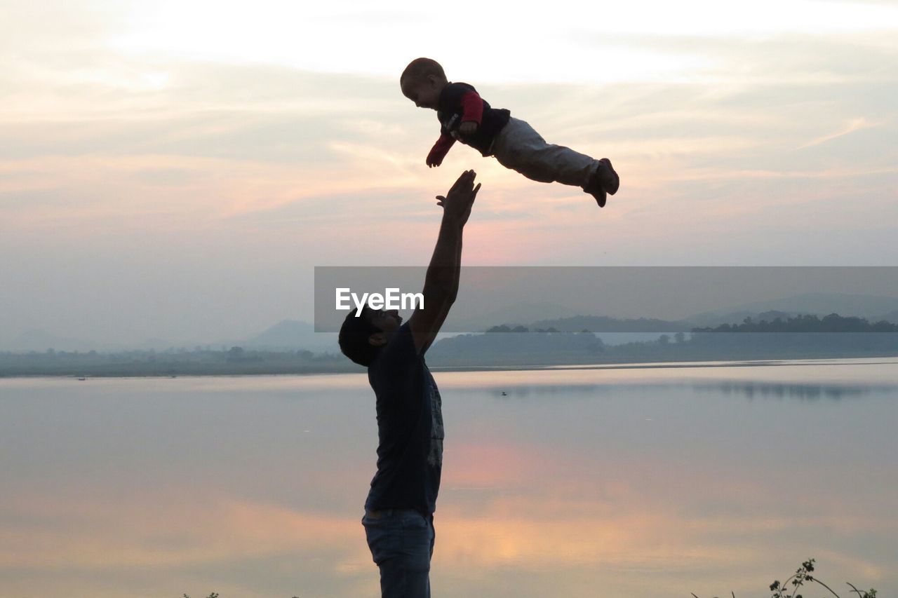 Father throwing son in air by lake against sky during sunset