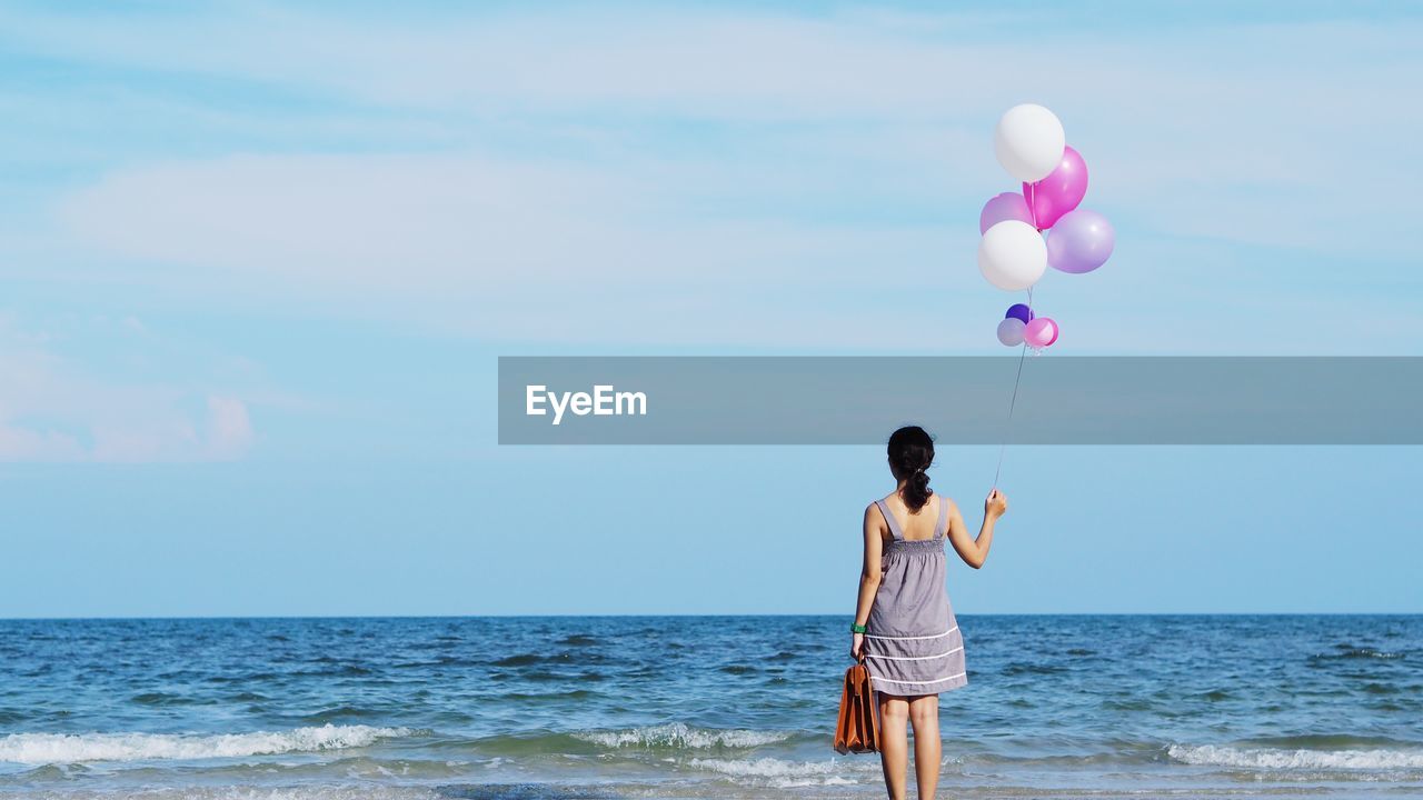 Rear view of woman with balloons on shore at beach against sky