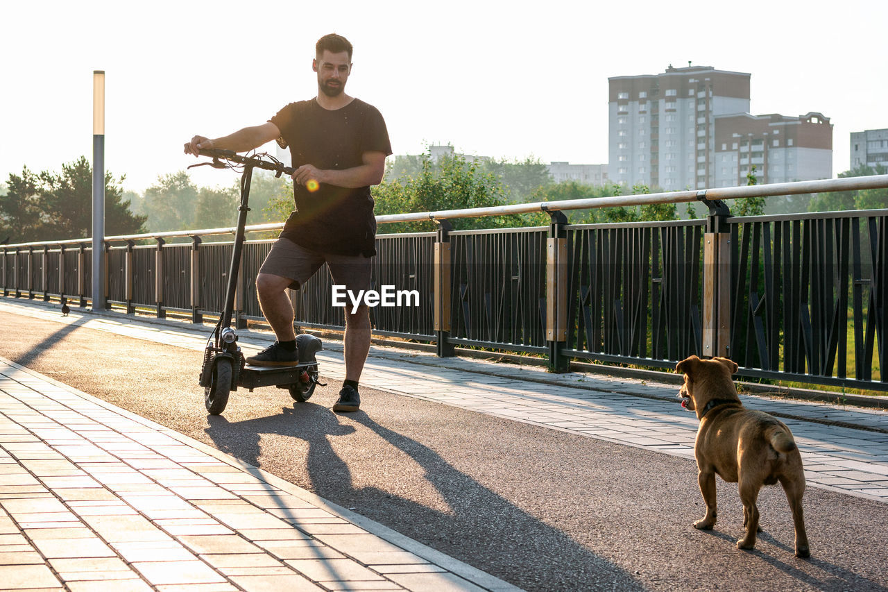 Handsome man is standing one leg on electric scooter and looking on his dog on the street. 