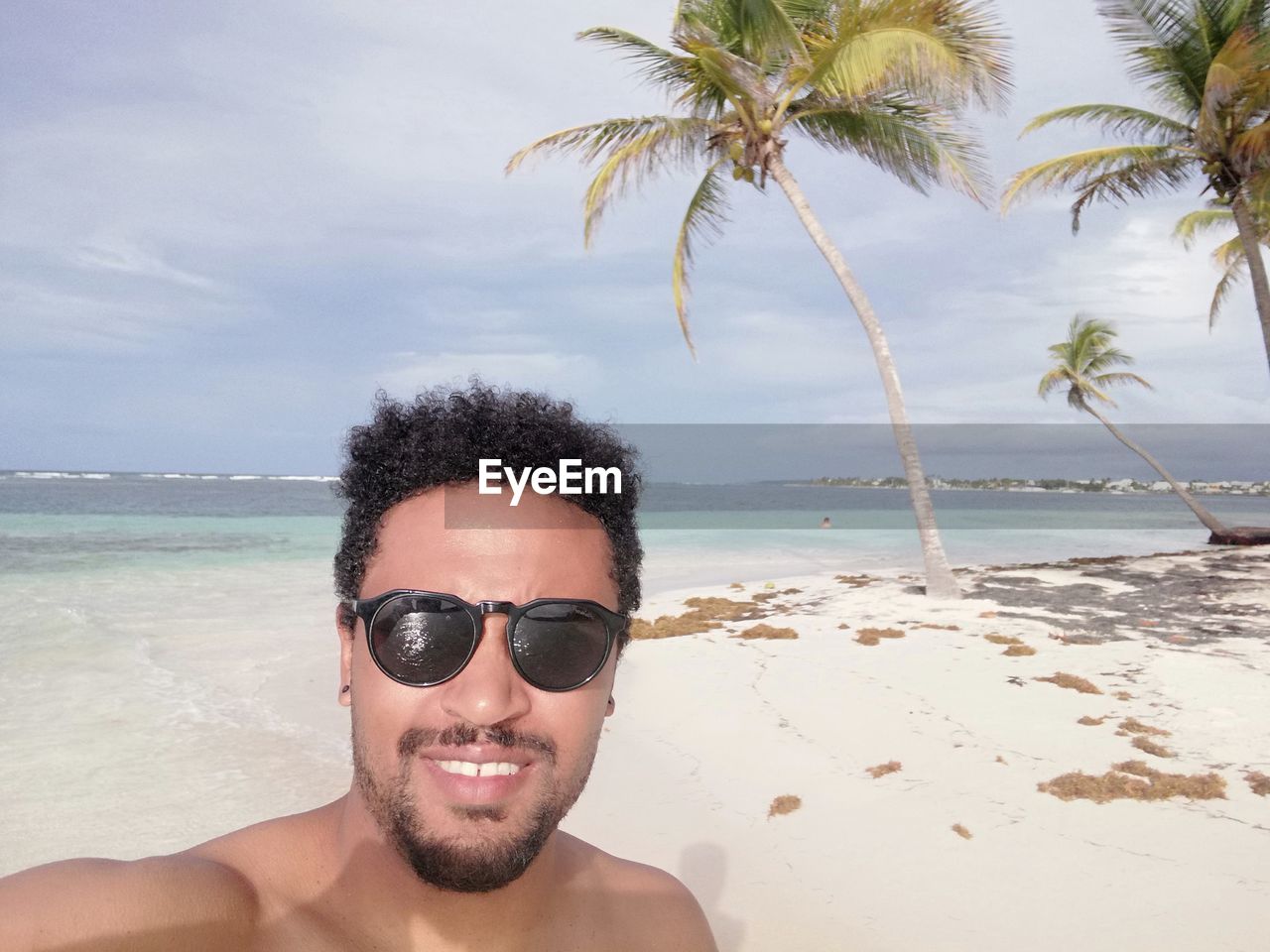 Portrait of smiling shirtless mid adult man wearing sunglasses standing at beach against cloudy sky