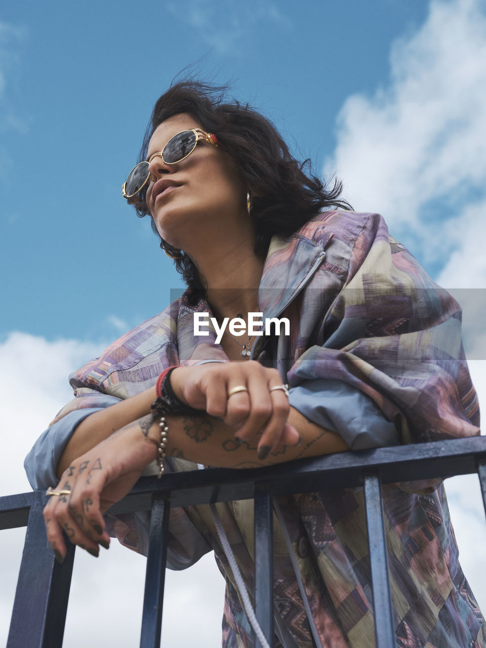 From below of trendy young female in sunglasses and with tattooed arms leaning on metal fence and looking away