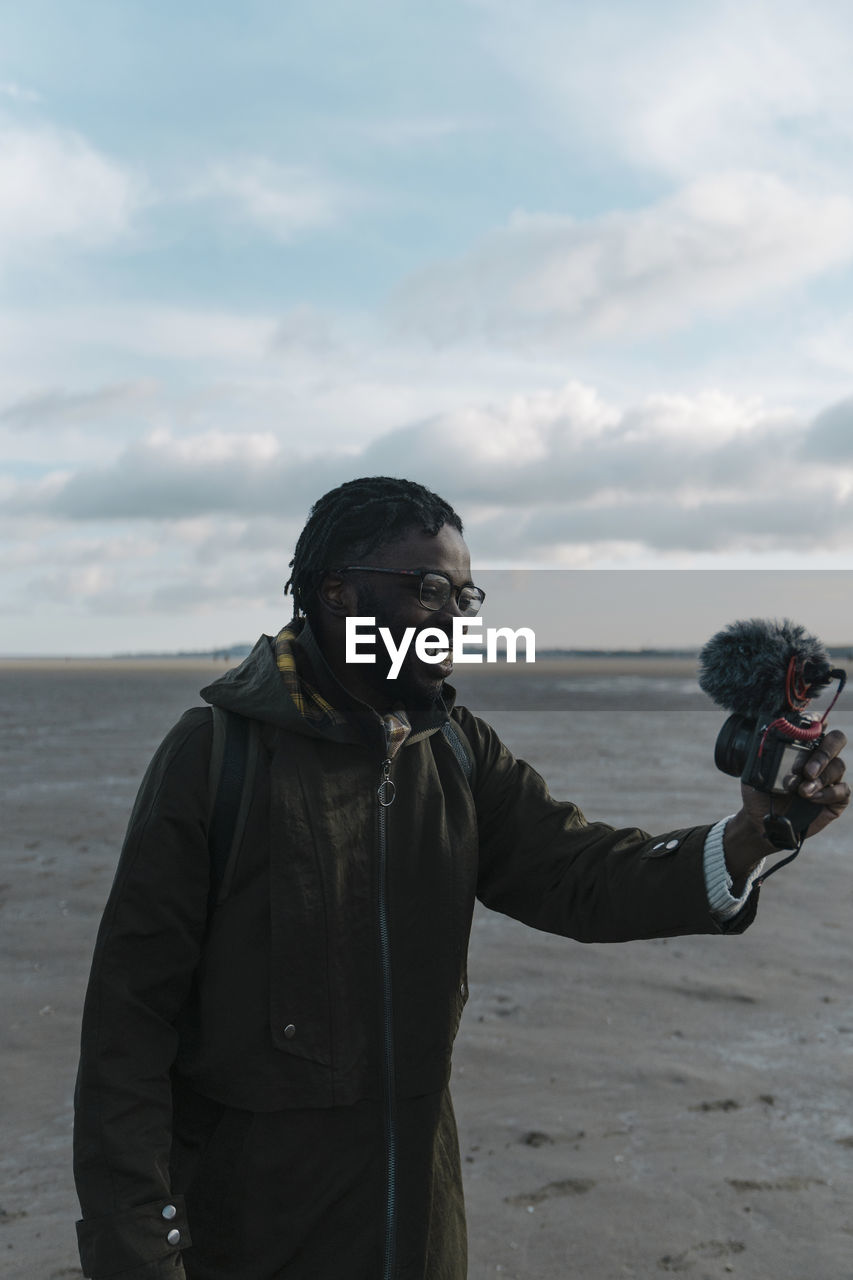 Smiling male vlogger live streaming through camera at beach against sky