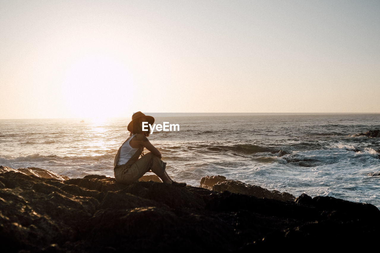 rear view of woman sitting on rock at beach against clear sky