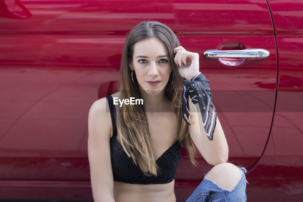 Portrait of young woman sitting on the street smiling looking at camera next to a red car