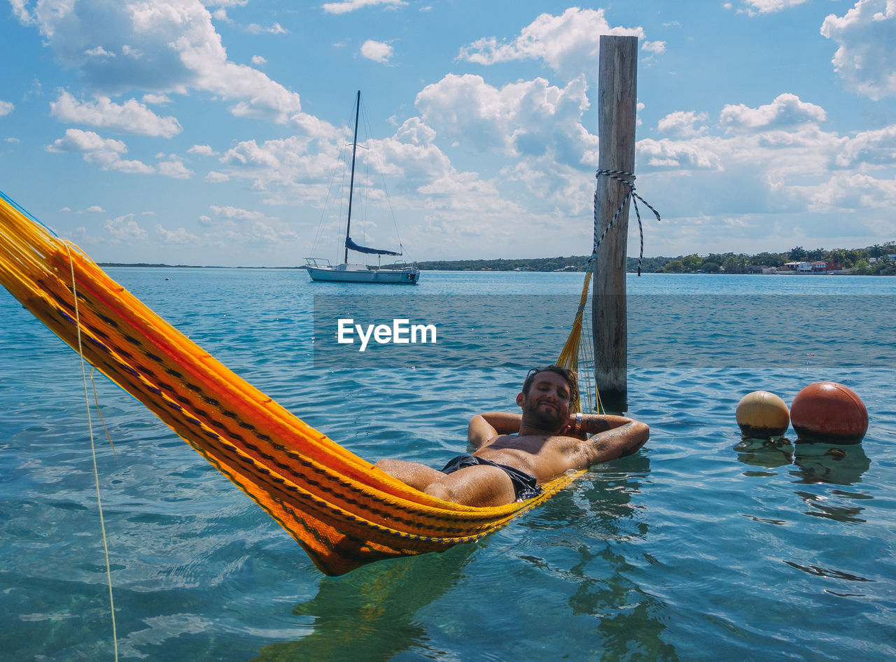 Shirtless young man lying on hammock in sea against cloudy sky during sunny day