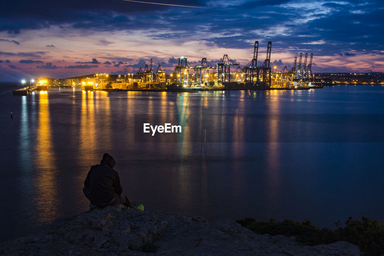 Man sitting on illuminated harbor against sky at dusk
