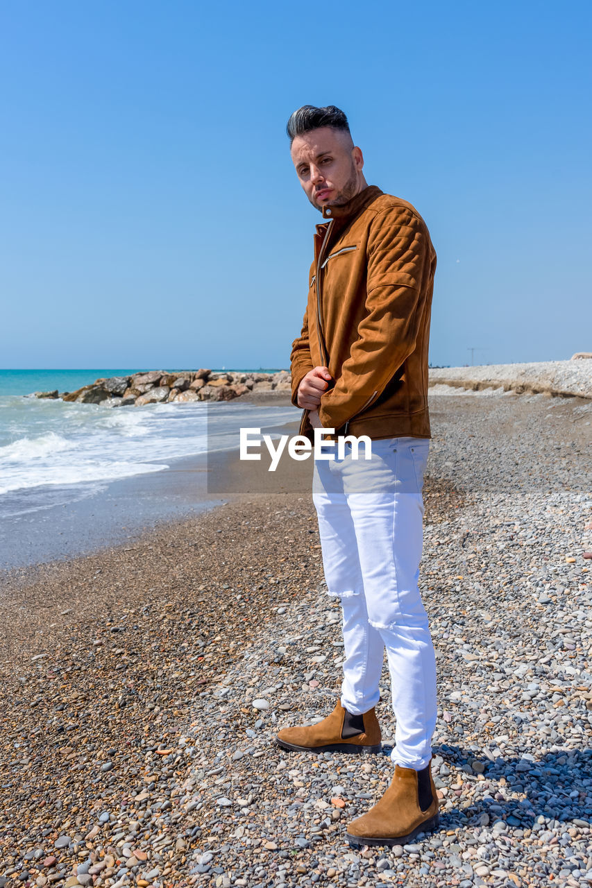 YOUNG MAN STANDING ON BEACH AGAINST CLEAR SKY