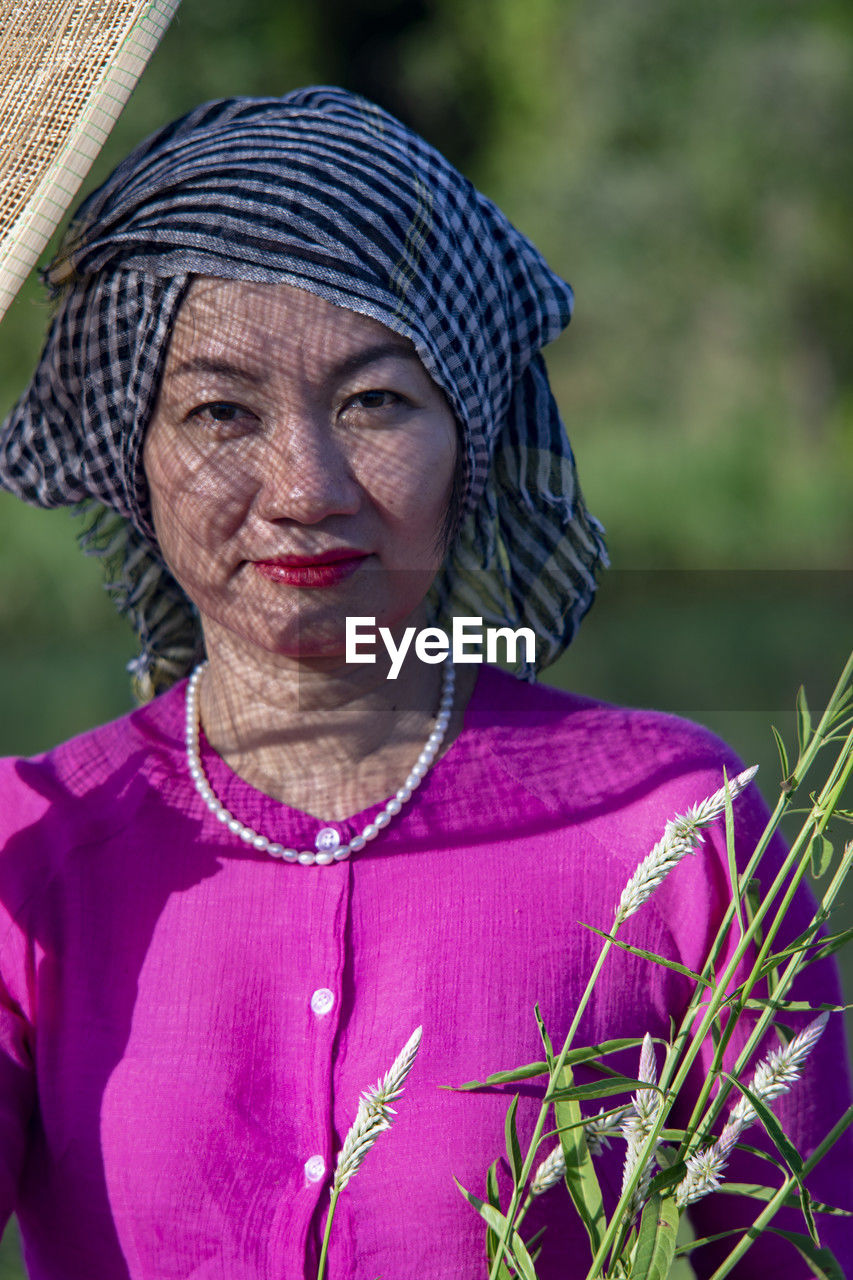 one person, adult, women, portrait, clothing, person, female, purple, plant, nature, front view, smiling, headshot, human face, lifestyles, looking at camera, emotion, outdoors, traditional clothing, senior adult, flower, rural scene, happiness, pink, mature adult, jewelry, landscape, waist up, day, focus on foreground, hat