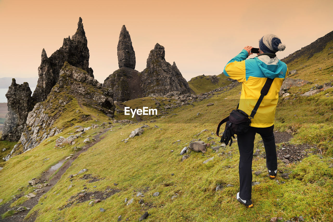 Rear view of man photographing old man of storr against sky during sunset