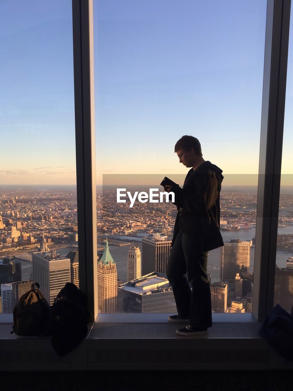 Man using mobile phone while standing on window sill during sunset