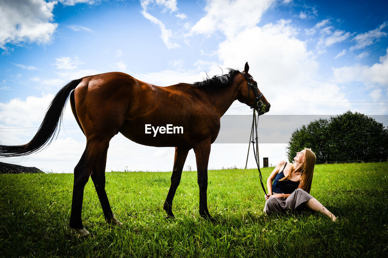 Smiling woman looking at horse while sitting on field against sky