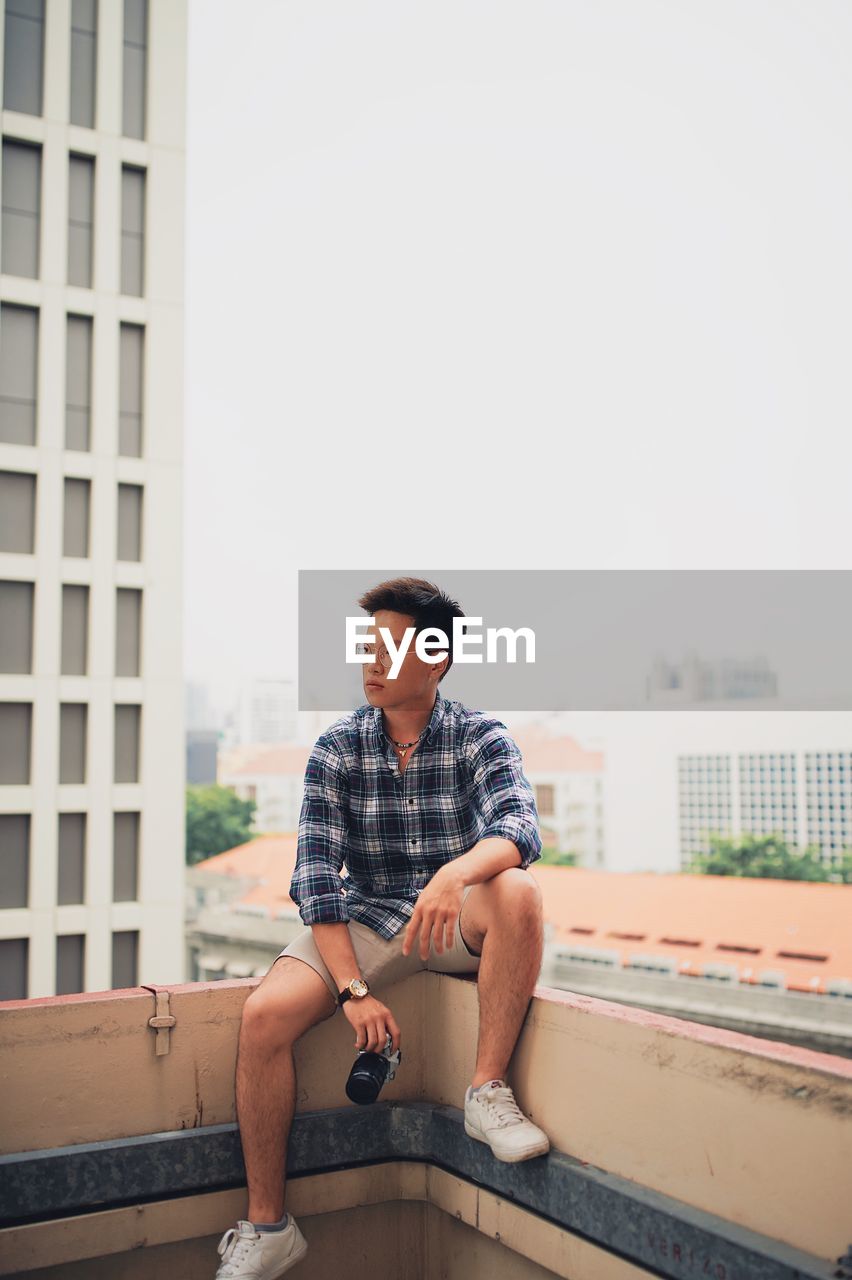 Young man with camera sitting on railing against clear sky