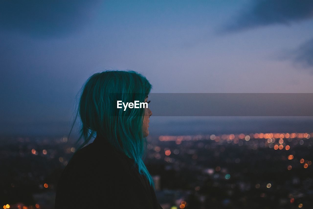 Side view of thoughtful woman standing against illuminated city at dusk