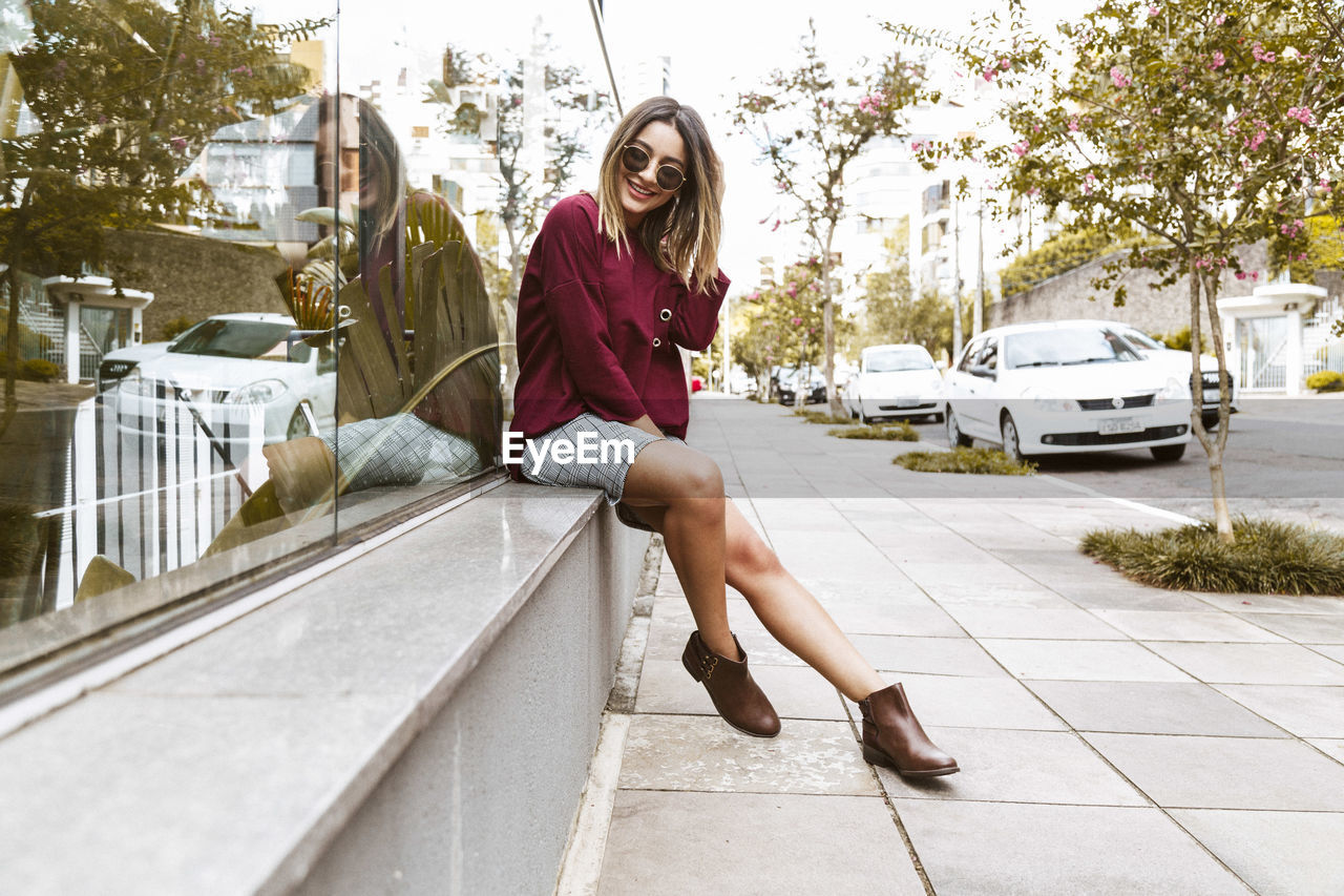 Portrait of smiling young woman sitting on sidewalk by glass building