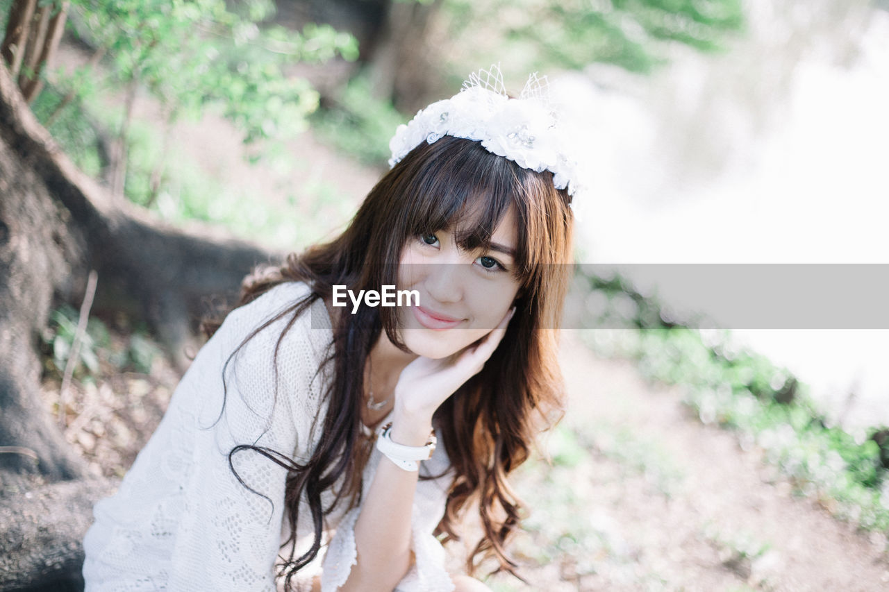 Portrait of beautiful young woman wearing white dress and wreath sitting on tree root in forest