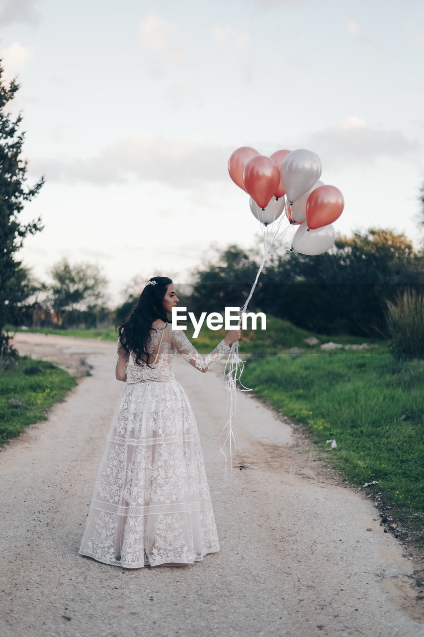 REAR VIEW OF WOMAN WITH BALLOONS STANDING ON ROAD