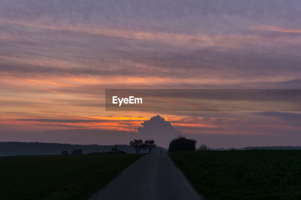 Road passing through field against cloudy sky