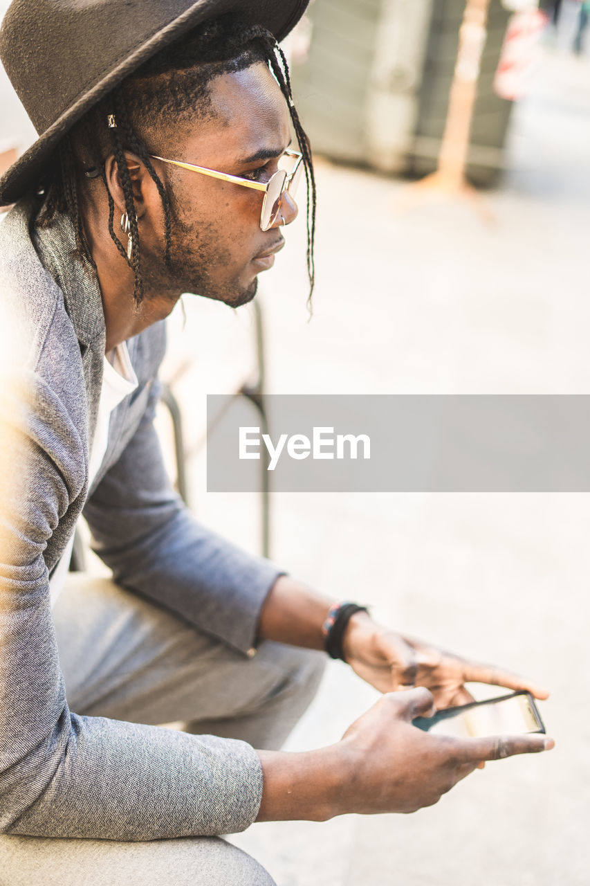 Side view of young man wearing sunglasses using mobile phone while sitting outdoors