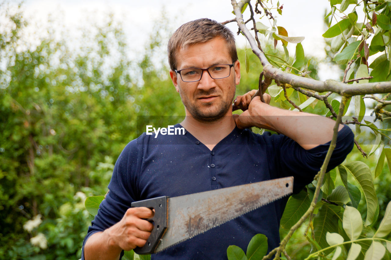 Portrait of man holding hand saw while standing against tree