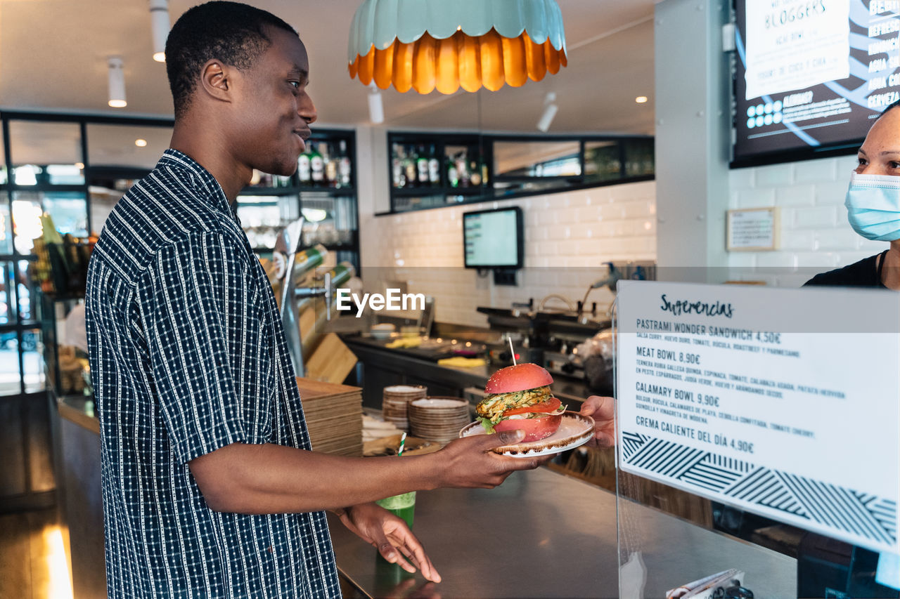 Side view of content black male with tasty burger on plate looking forward against signboard in restaurant