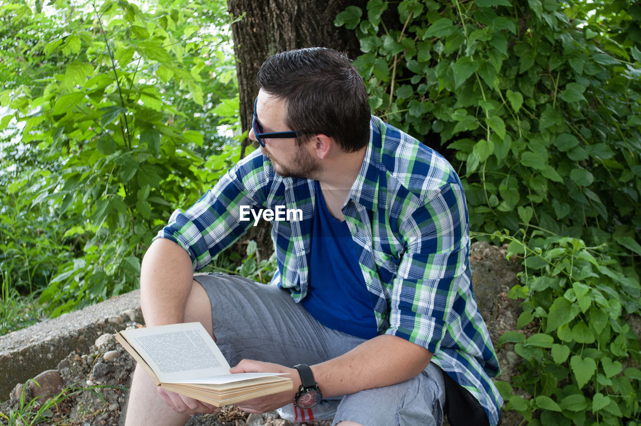 Young man looking away while holding book in forest