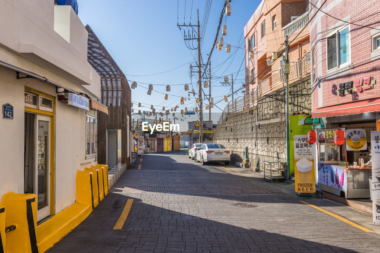 architecture, road, city, neighbourhood, street, building exterior, town, urban area, built structure, infrastructure, transportation, lane, city street, building, sky, mode of transportation, city life, downtown, nature, travel destinations, day, transport, sign, residential district, outdoors, residential area, vehicle, travel, sunlight, sunny, footpath, car, alley, yellow, store, no people, motor vehicle, cityscape, clear sky