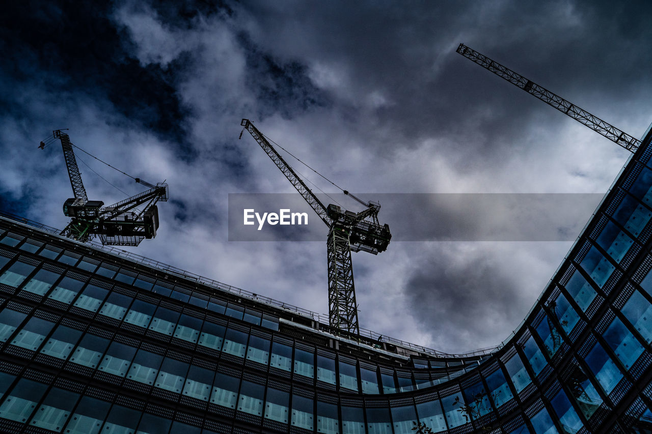 Low angle view of cranes on building against cloudy sky