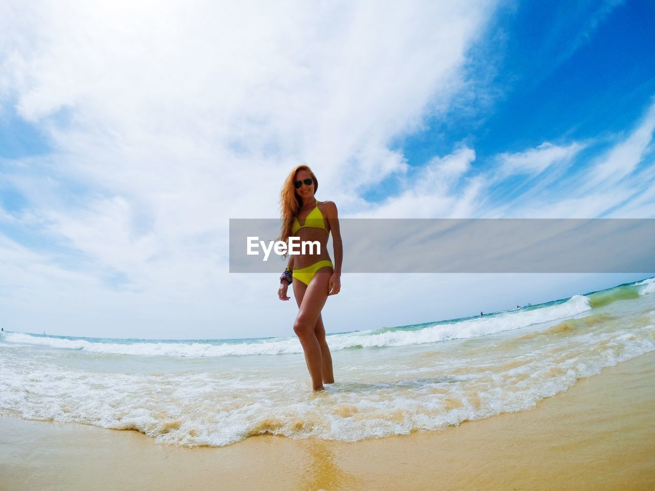 Low angle view of mid adult woman in bikini standing at beach against cloudy sky