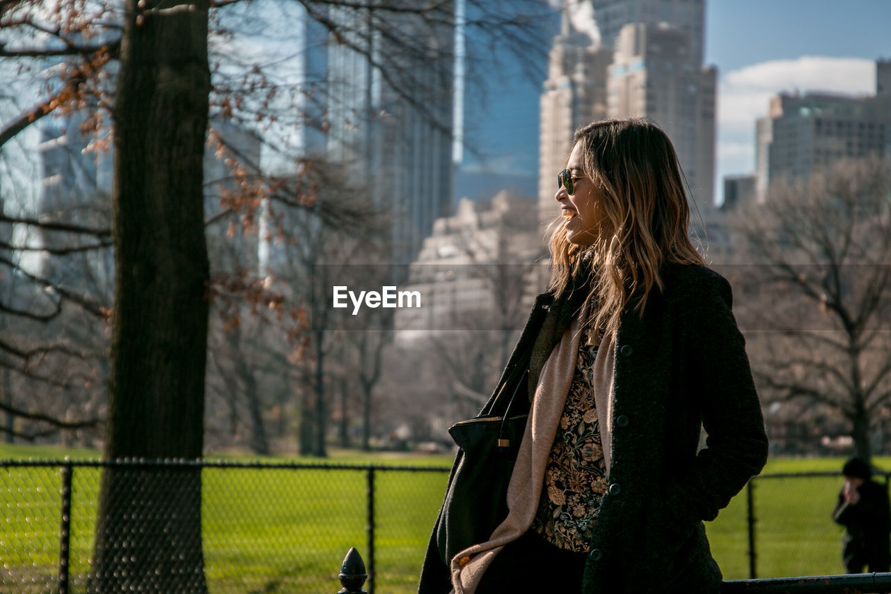 Happy woman wearing jacket standing by trees and buildings