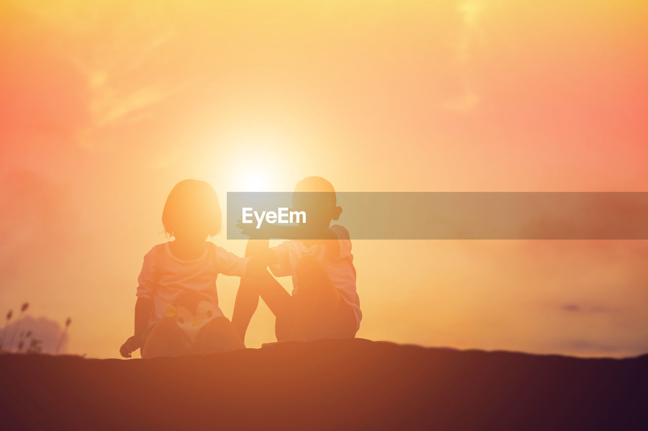 Siblings sitting on land against sky during sunset