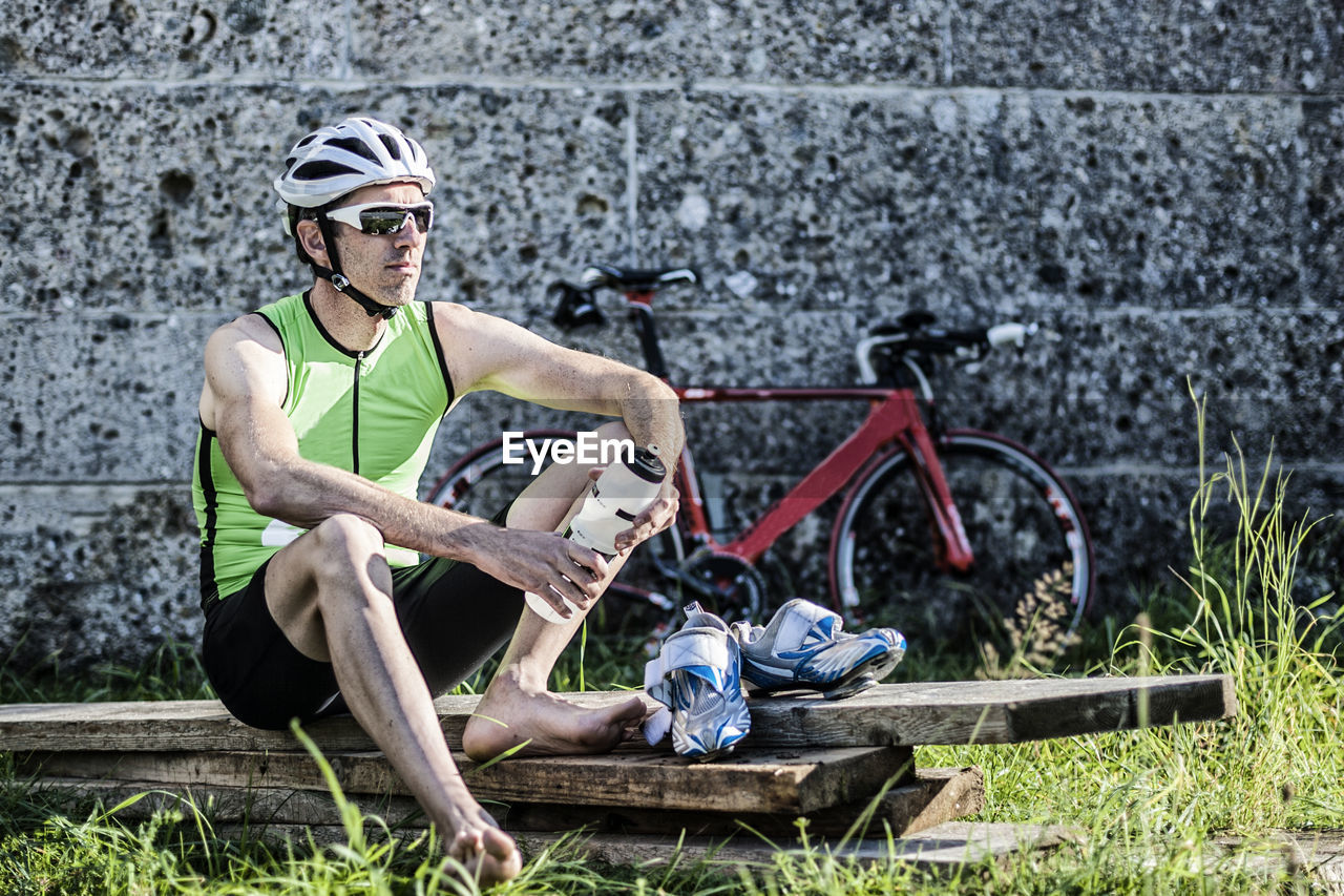 Cyclist holding water bottle while relaxing on wooden plank against wall