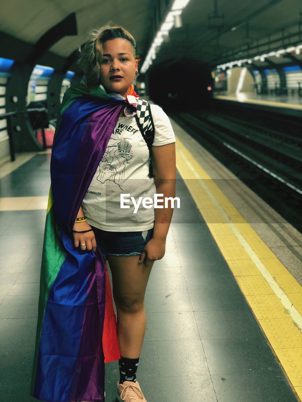 Portrait of young woman standing with rainbow flag at railroad station platform