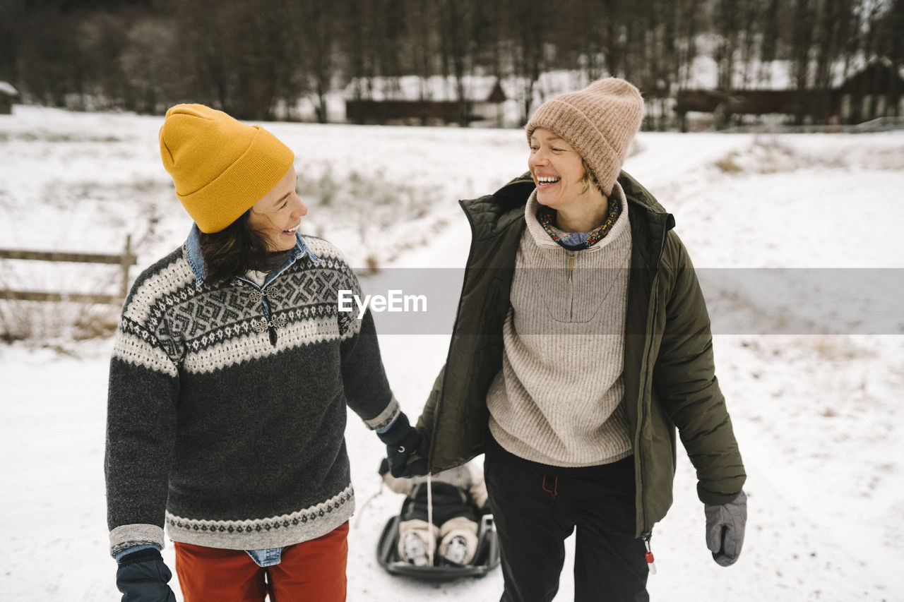 Smiling lesbian couple looking at each other while pulling daughter on snow