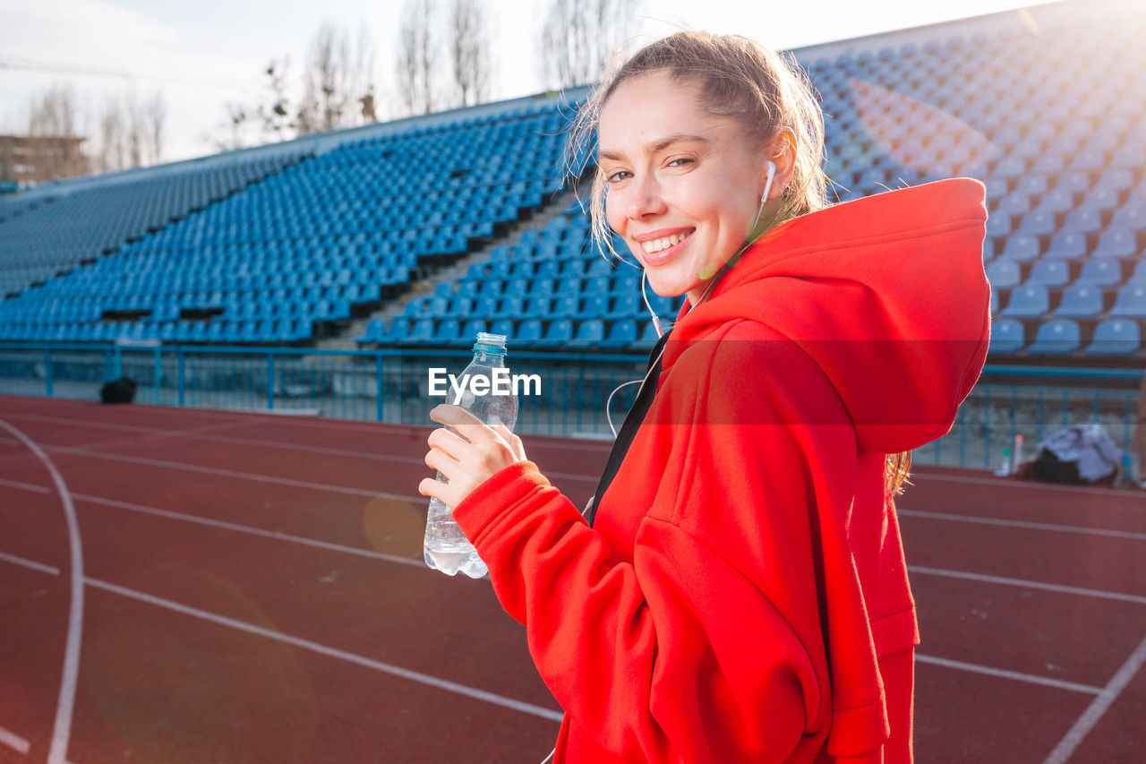 Portrait of smiling woman drinking water while listening music on running track