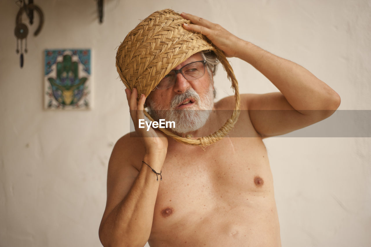 Shirtless man with basket on head standing against wall at home