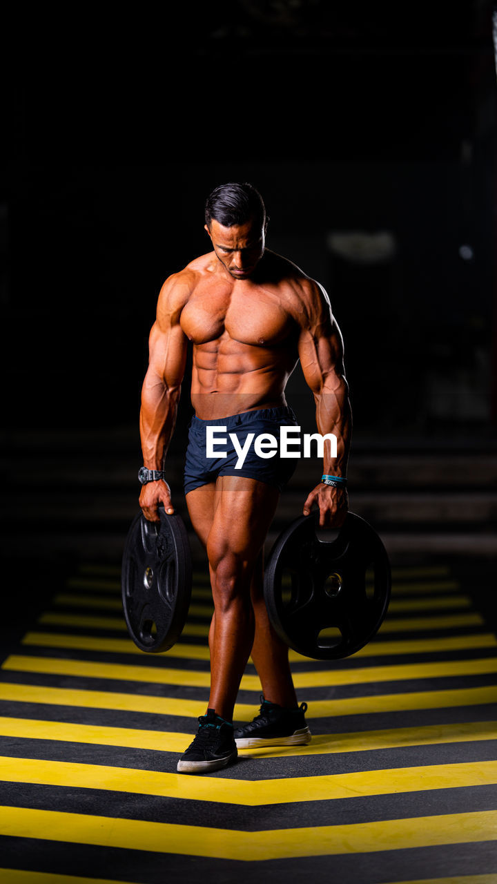 Full length of shirtless man holding weights while walking on floor