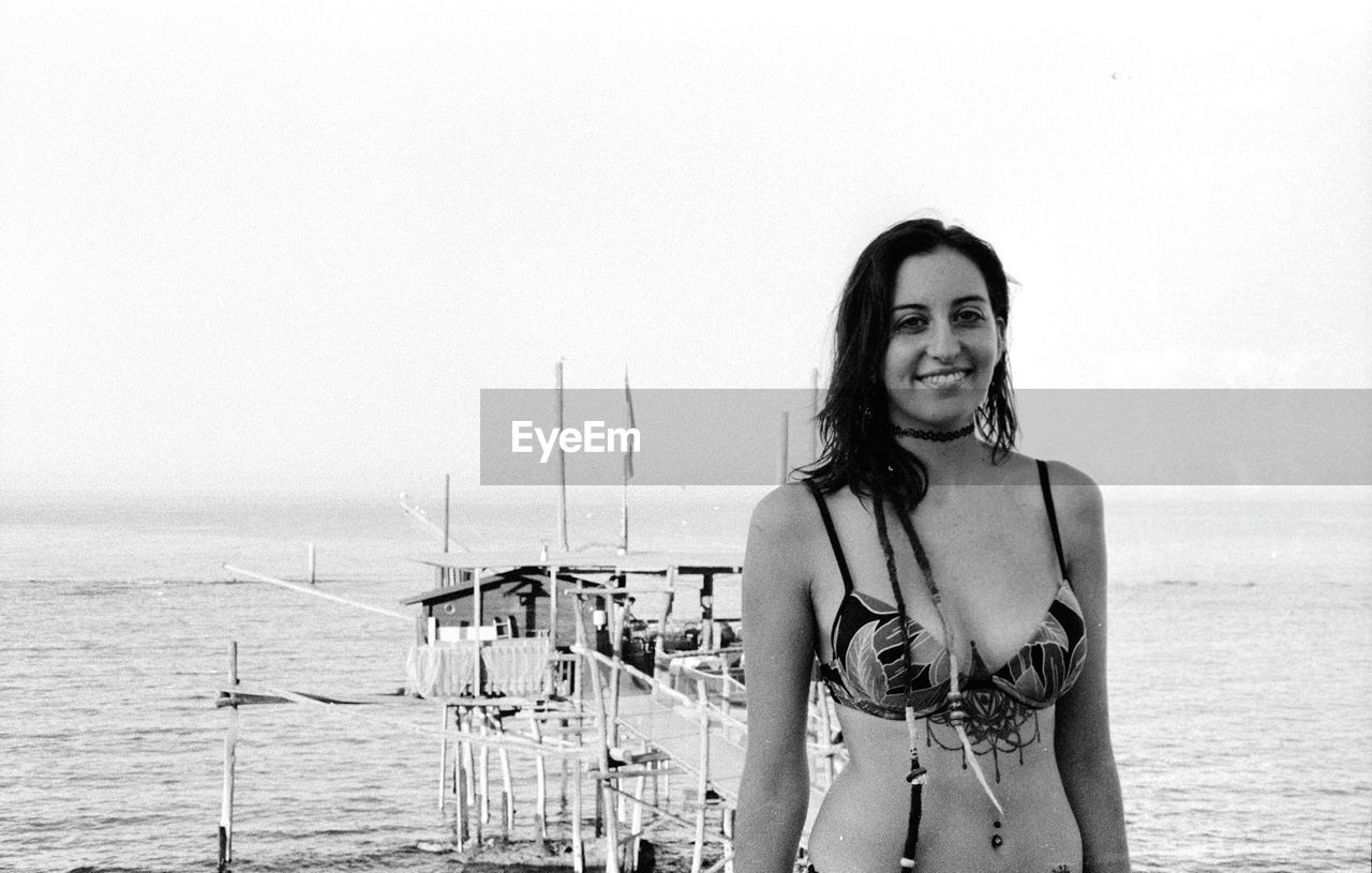 Portrait of smiling young woman standing at beach against clear sky