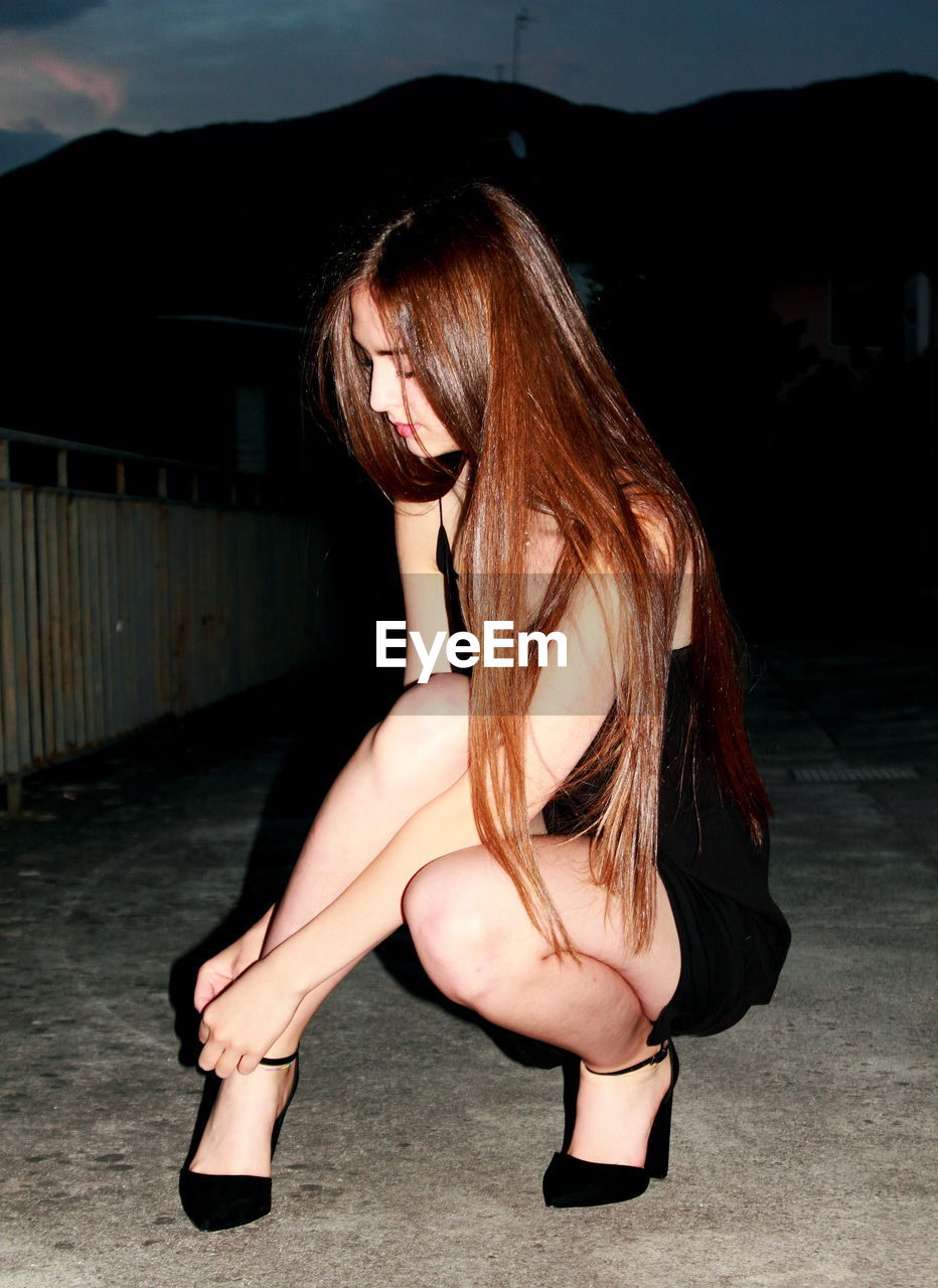 Young woman with long brown hair wearing high heels on sidewalk at night