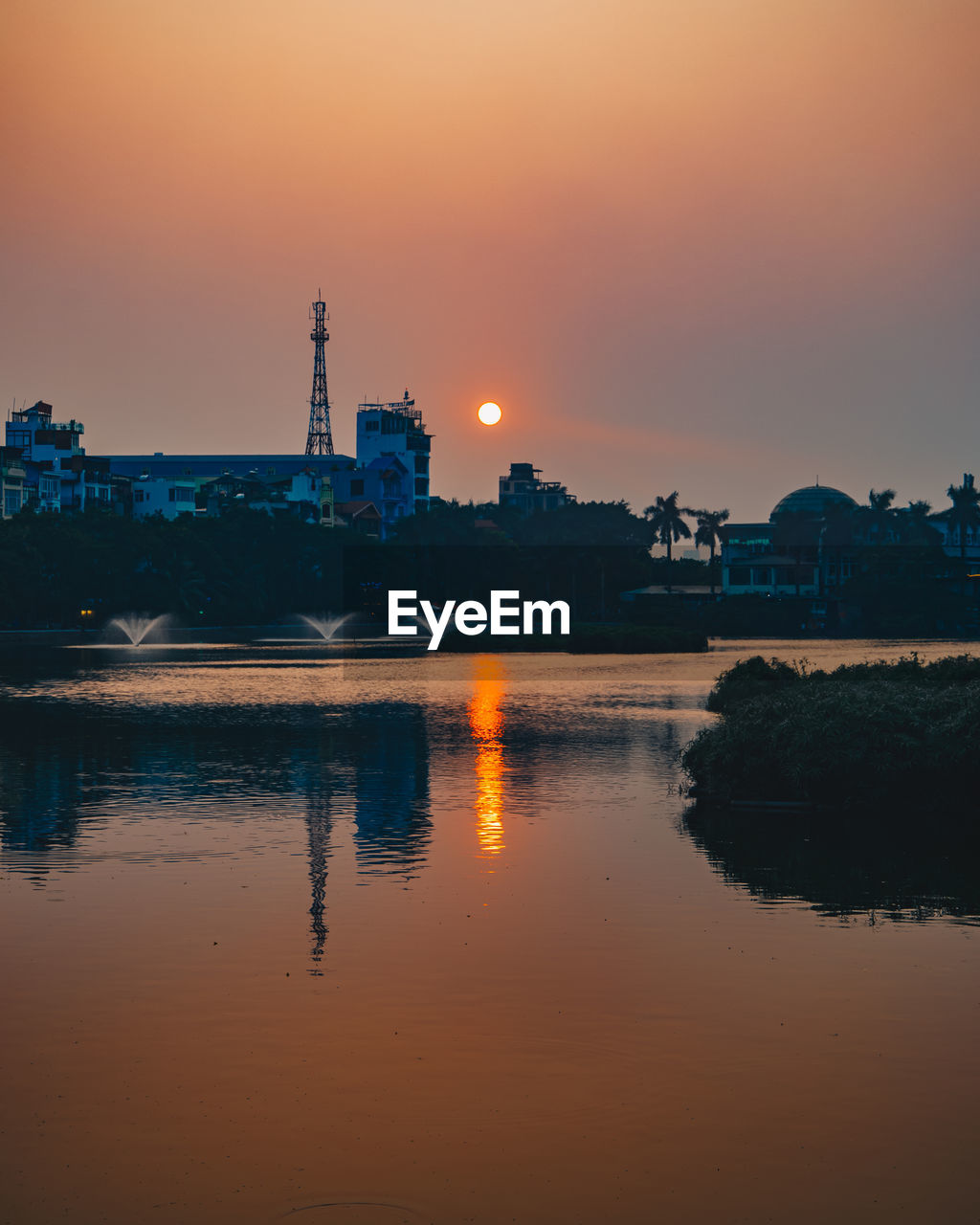 River by illuminated factory against sky during sunset