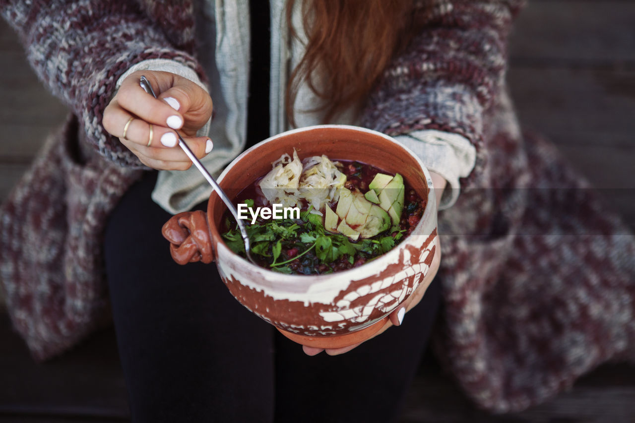 Midsection of woman holding food in bowl with spoon while sitting outdoors