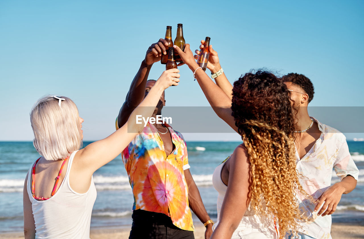 Cheerful young diverse best friends smiling and toasting with bottles of beer against cloudless blue sky on sunny summer day