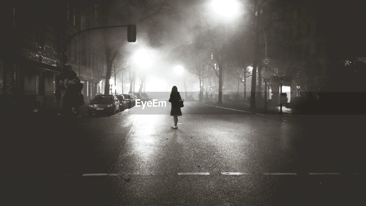 LOW ANGLE VIEW OF WOMAN STANDING ON ILLUMINATED STREET AT NIGHT
