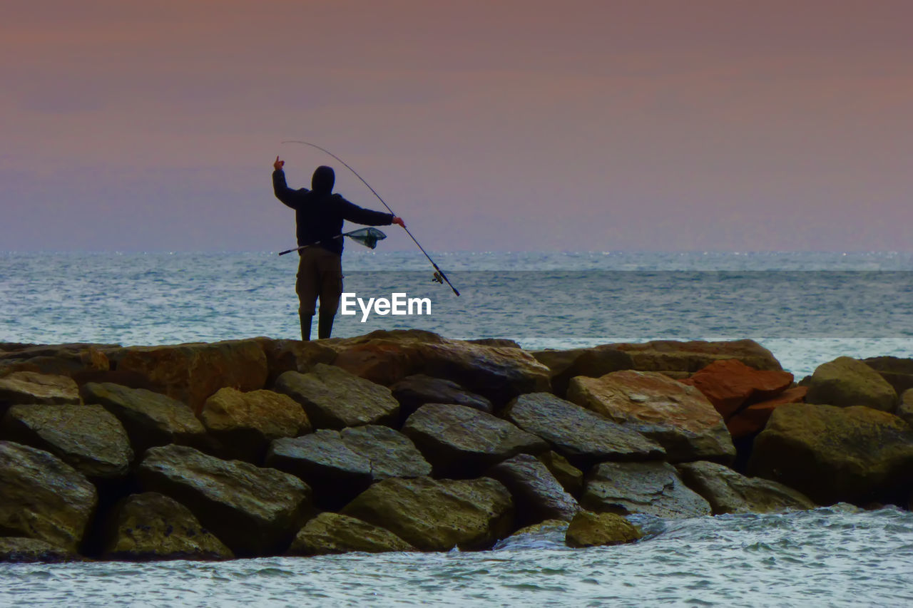 FULL LENGTH OF SILHOUETTE MAN STANDING ON ROCK AGAINST SEA