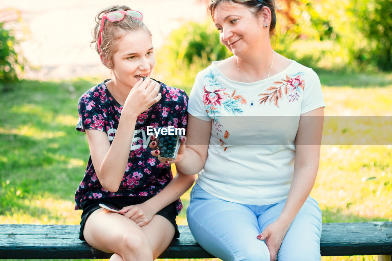 Smiling mature woman holding blueberries in glass by daughter sitting on bench