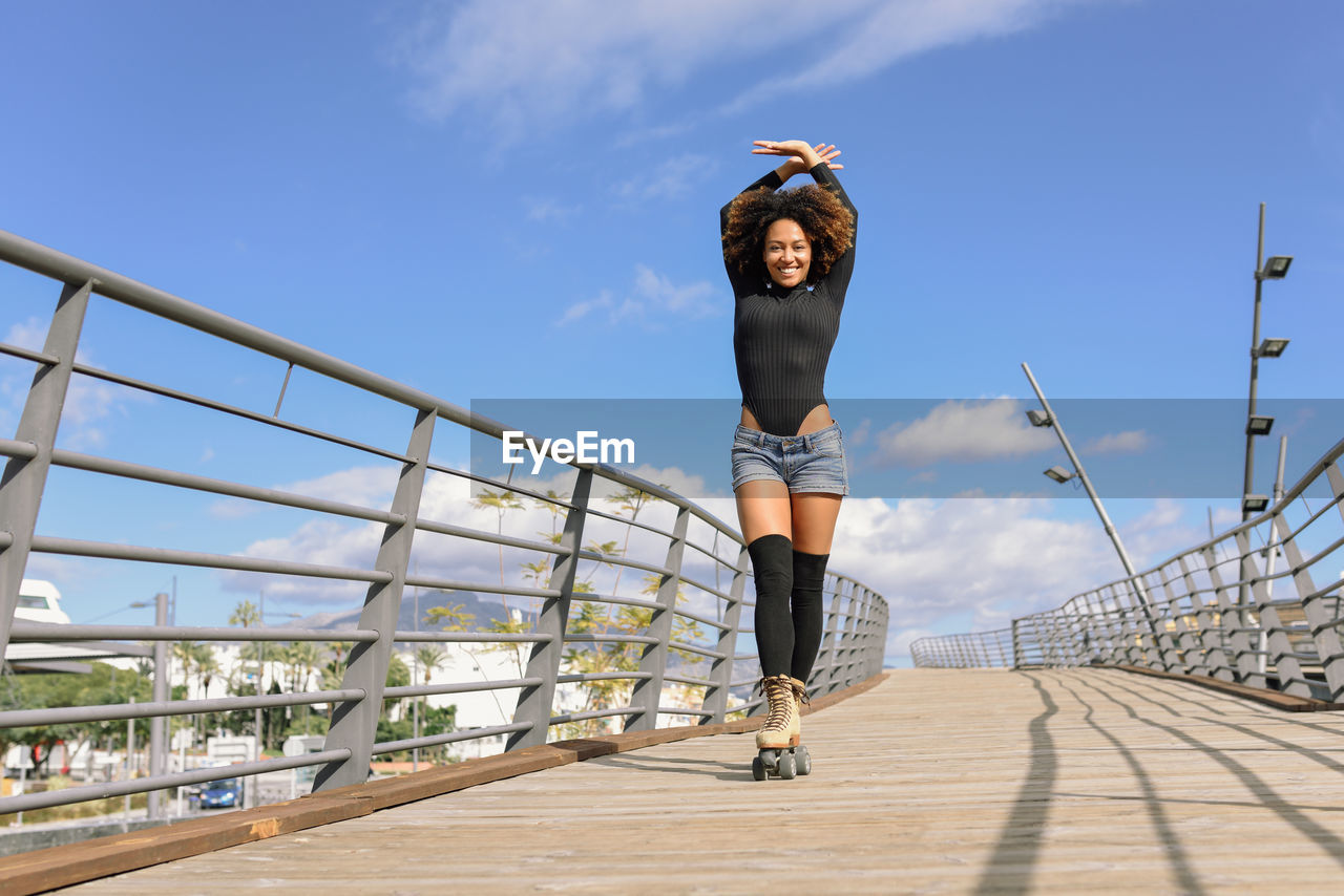 Portrait of woman with arms raised wearing roller skates on footbridge in city against sky