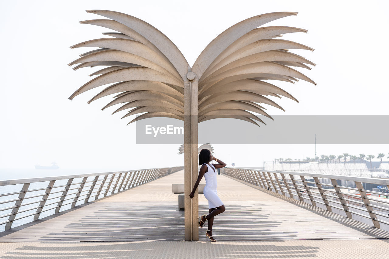 Attractive african american female elegant white dress standing on embankment near wing shaped structure and looking away