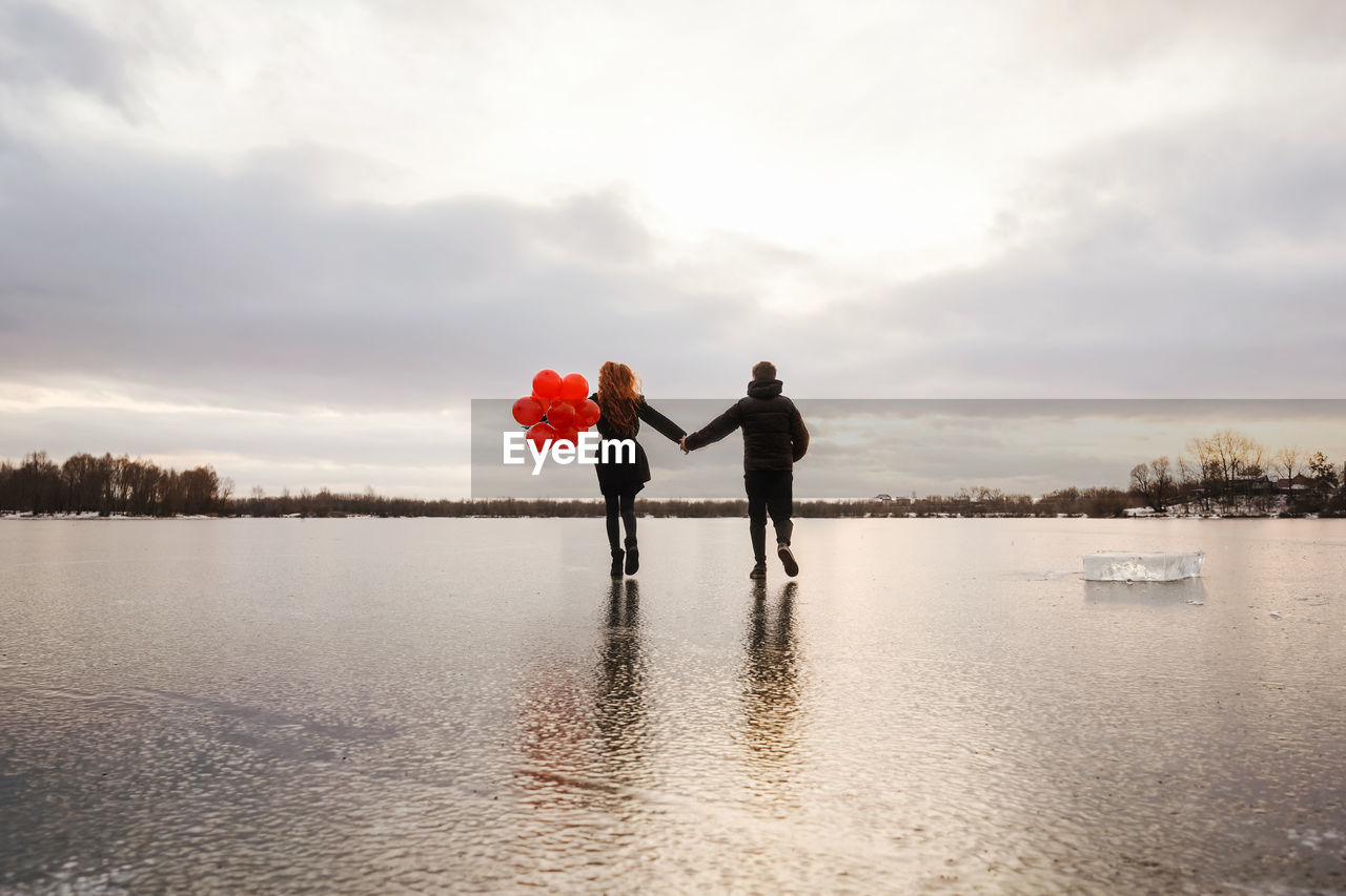 Rear view of couple holding hand walking on frozen lake against sky
